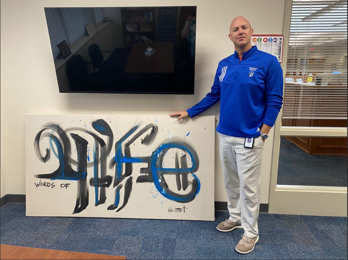 East principal Casey Fries standing in his office next to a painting done by Eric Timm during Timms hope assembly, September 22, 2023. The painting is one of many done by Timm, and a recreation of a previous painting that used to reside in the school commons that he made for East during his last visit several years ago.
