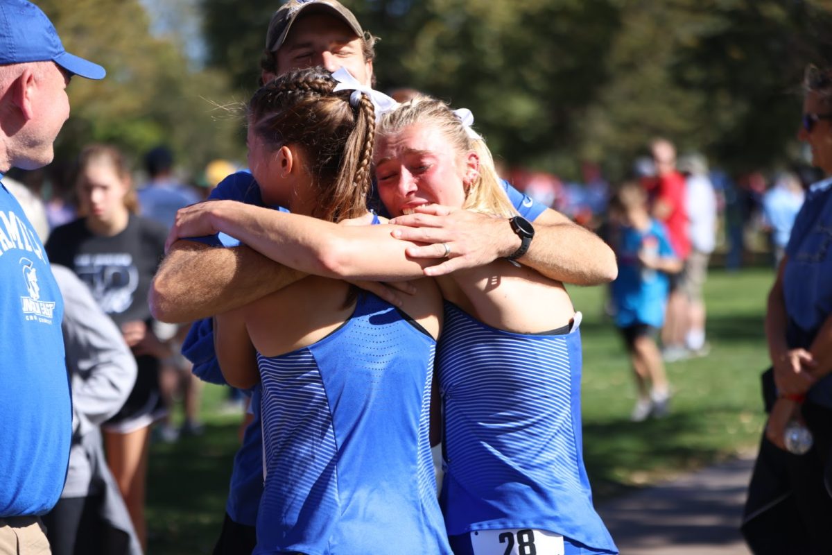 Senior Jordyn Wissing (L) and sophomore Ella Herzberg (R) embrace assistant coach Andrew Willis after the state cross country race on October 20, 2023. Head coach Brian Kabourek witnesses the embrace.